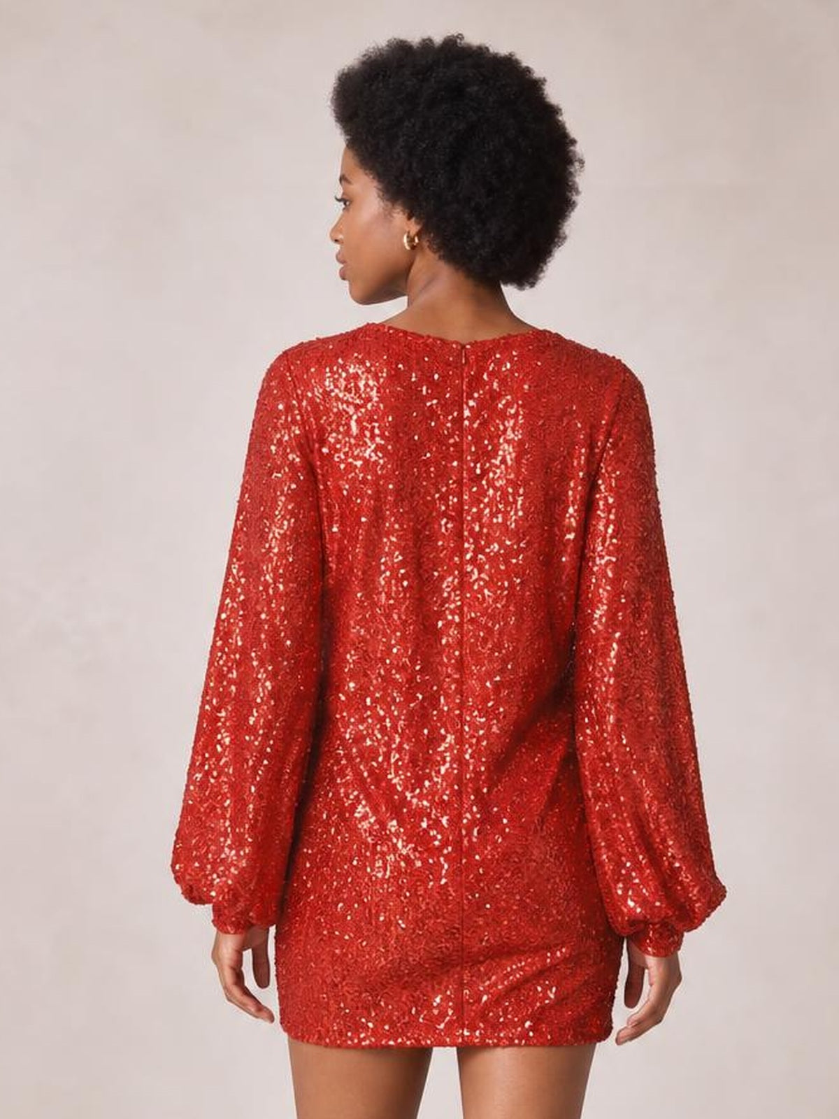 A model poses facing away in a red sequin long sleeve mini dress with a round neckline, straight relaxed silhouette, and visible center-back zipper.