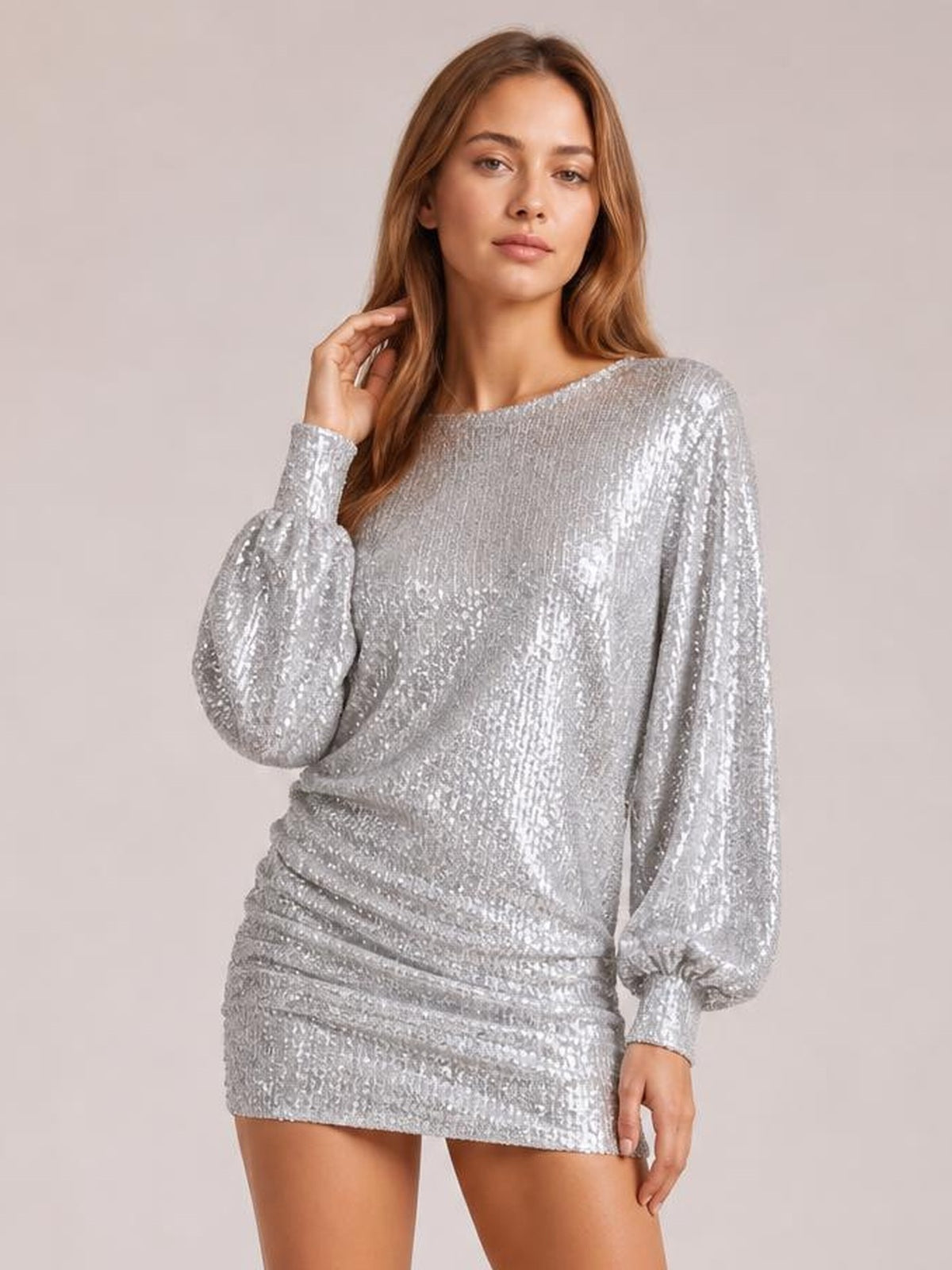 Model poses with one hand near her hair in a silver sequin long sleeve mini dress featuring a fitted silhouette, round neckline, and ruched skirt.