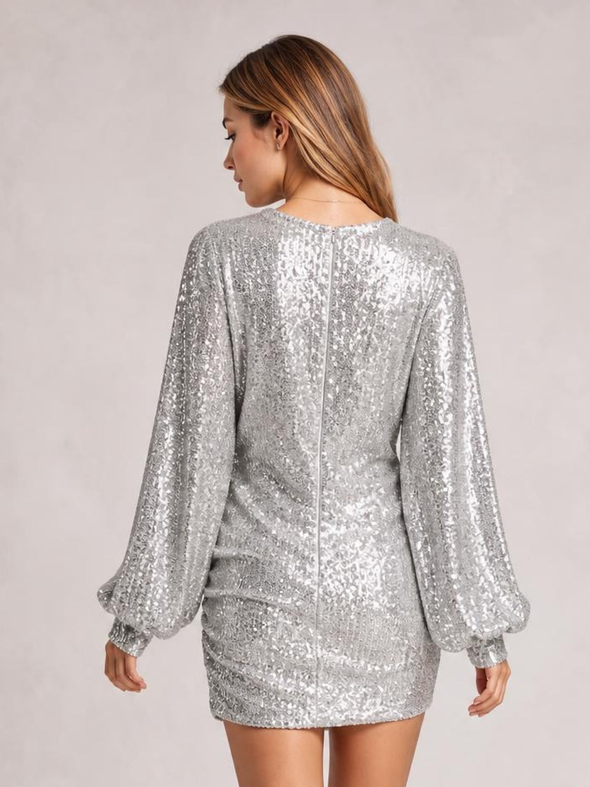 A model stands turned away in a silver sequin long sleeve ruched mini dress with voluminous sleeves, gathered waist, and a visible center-back zipper.
