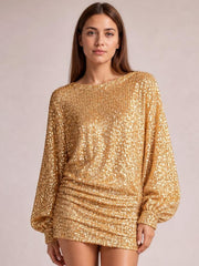 A model stands in a gold sequin long sleeve ruched mini dress with a round neckline, lined construction, and a relaxed silhouette.