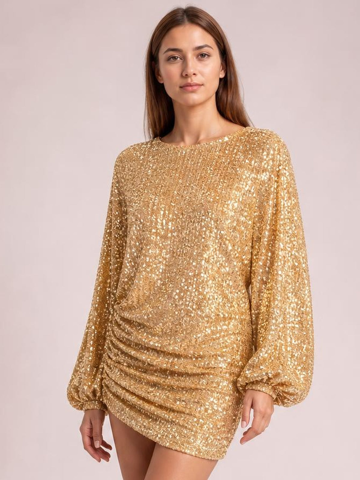 Model wears a gold sequin long-sleeve ruched mini dress with a round neckline, lined silhouette, and gathered detailing at the hips.