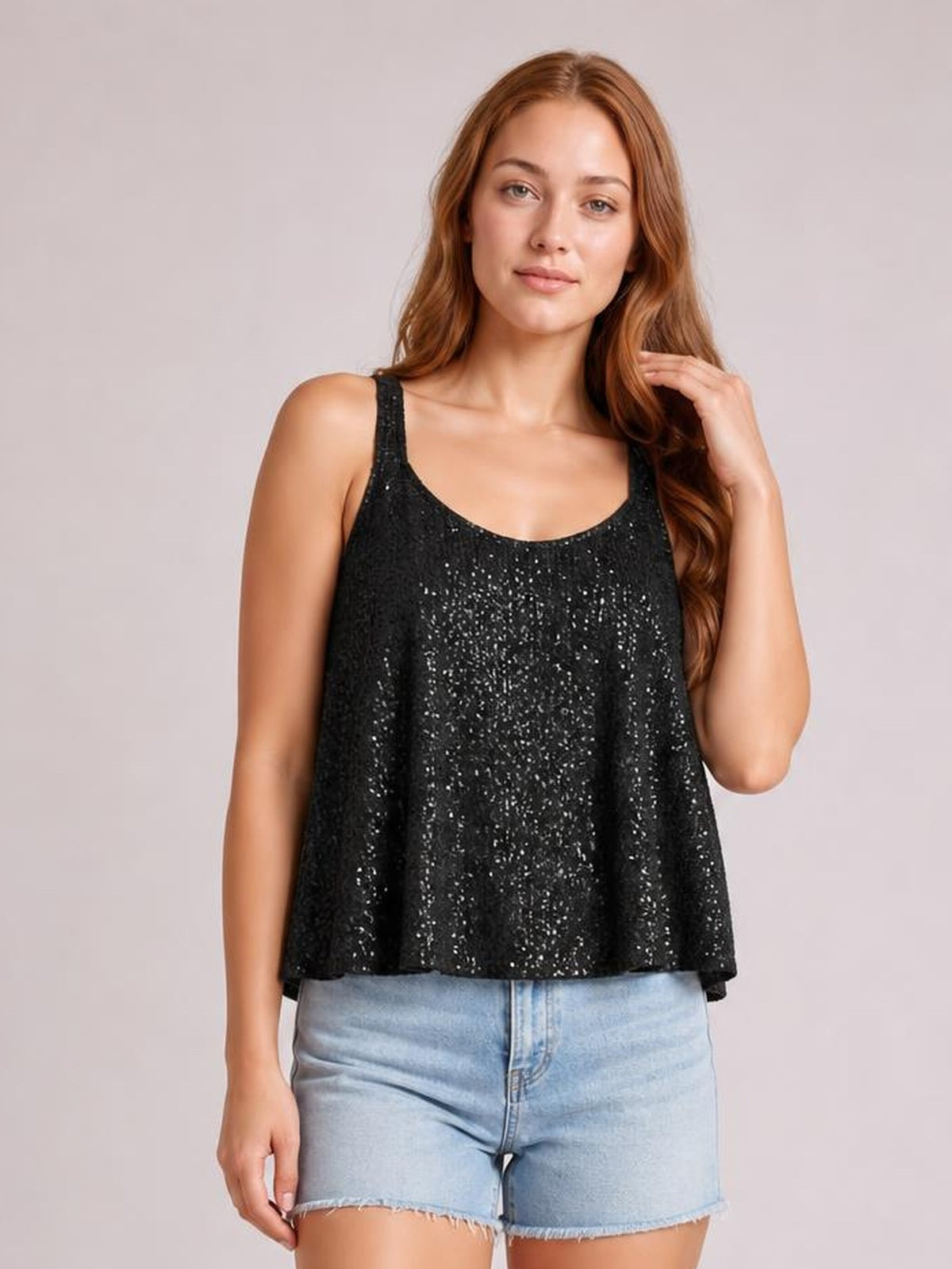 Model wearing a black sequin scoop neck tank with wide straps and a loose, subtly flared silhouette, paired with denim shorts while touching her hair.