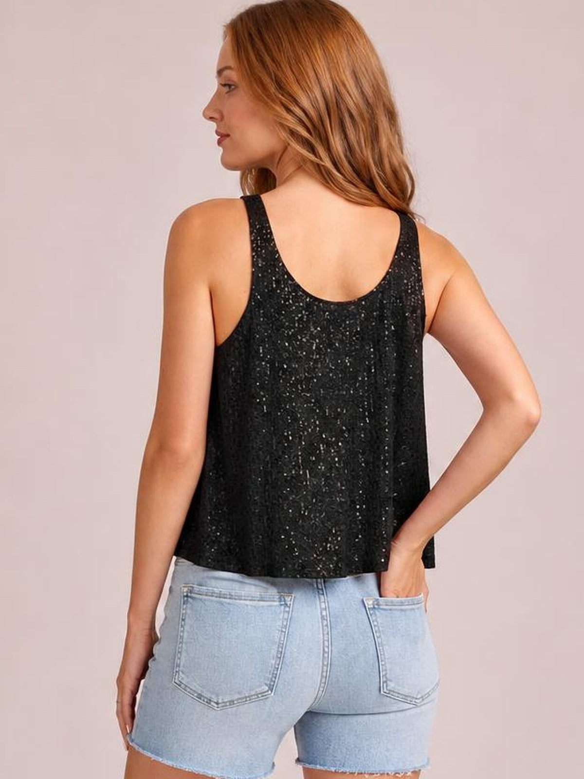A model turns to show the back of a black sequin scoop neck tank with wide straps and a relaxed straight silhouette, paired with light-wash denim shorts.