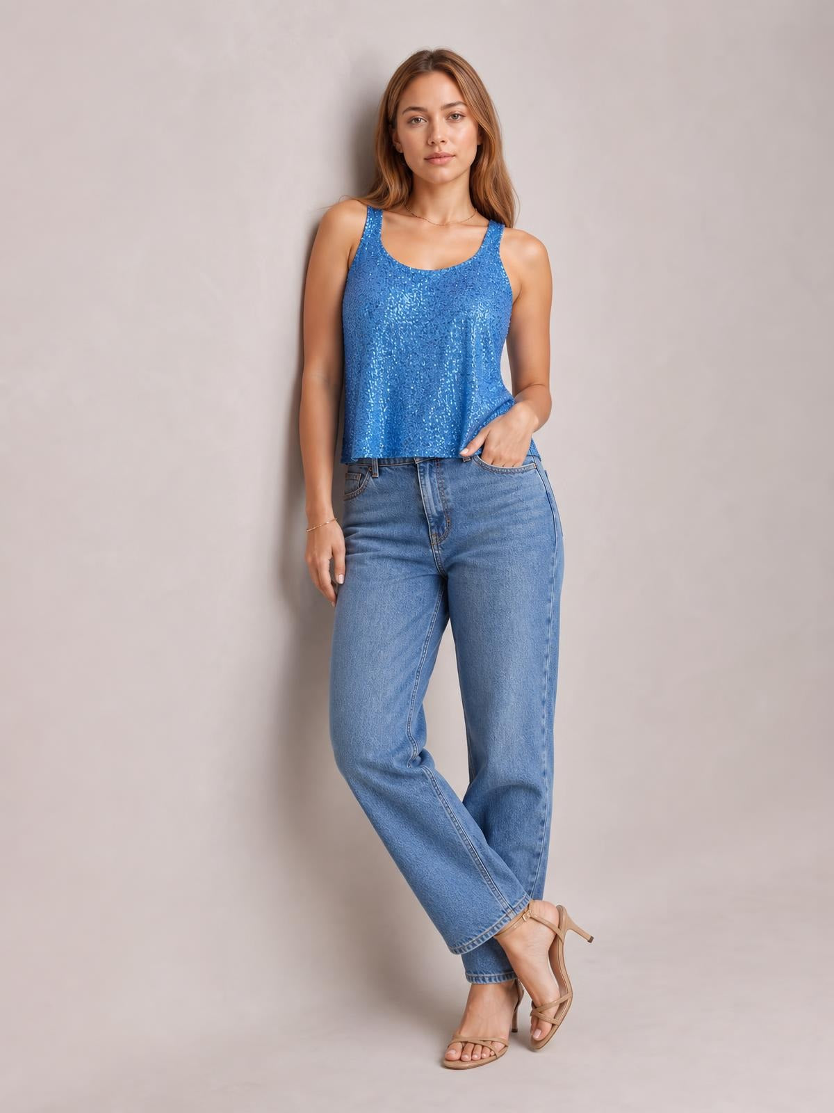 Model poses in a blue sequin scoop neck tank with wide straps and a relaxed straight silhouette, lightly holding the hem above her jeans.