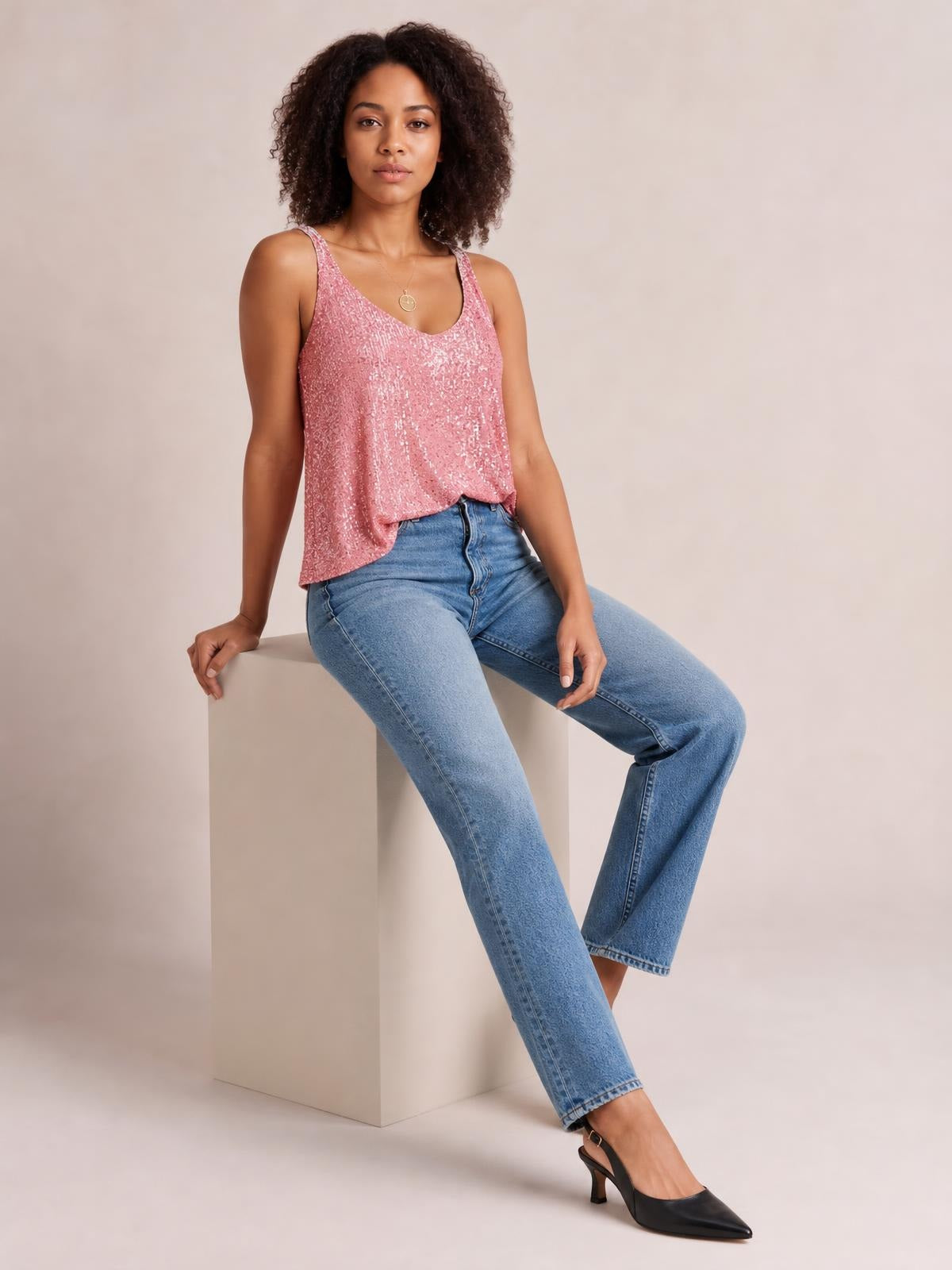 A model sits on a cube wearing a pink sequin scoop neck tank with thin straps and a relaxed straight silhouette, styled with light-wash jeans.
