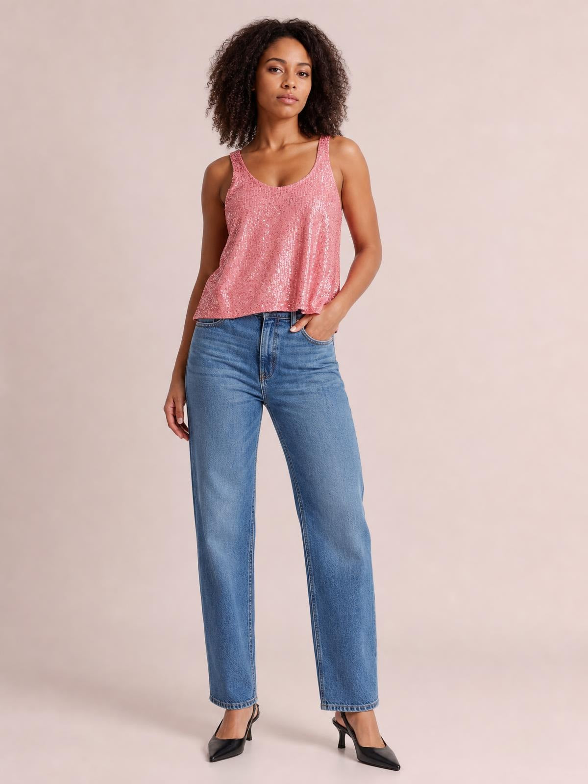 Model wears a pink sequin scoop neck tank with wide shoulder straps and a relaxed, slightly flared silhouette, standing with one hand in her jeans pocket.