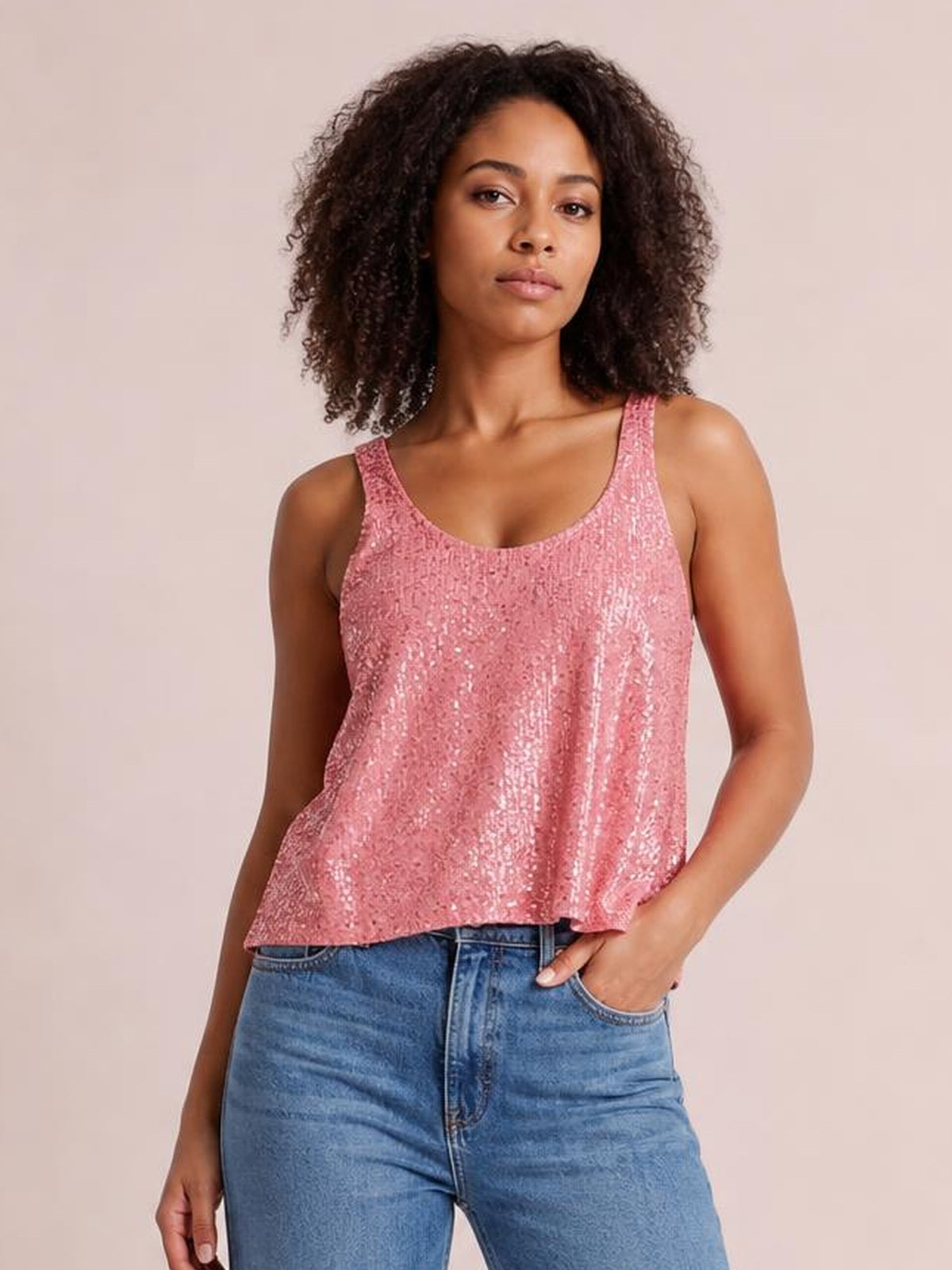 A model poses in a pink sequin scoop neck tank with wide shoulder straps and a relaxed, slightly flared silhouette, paired with blue jeans.