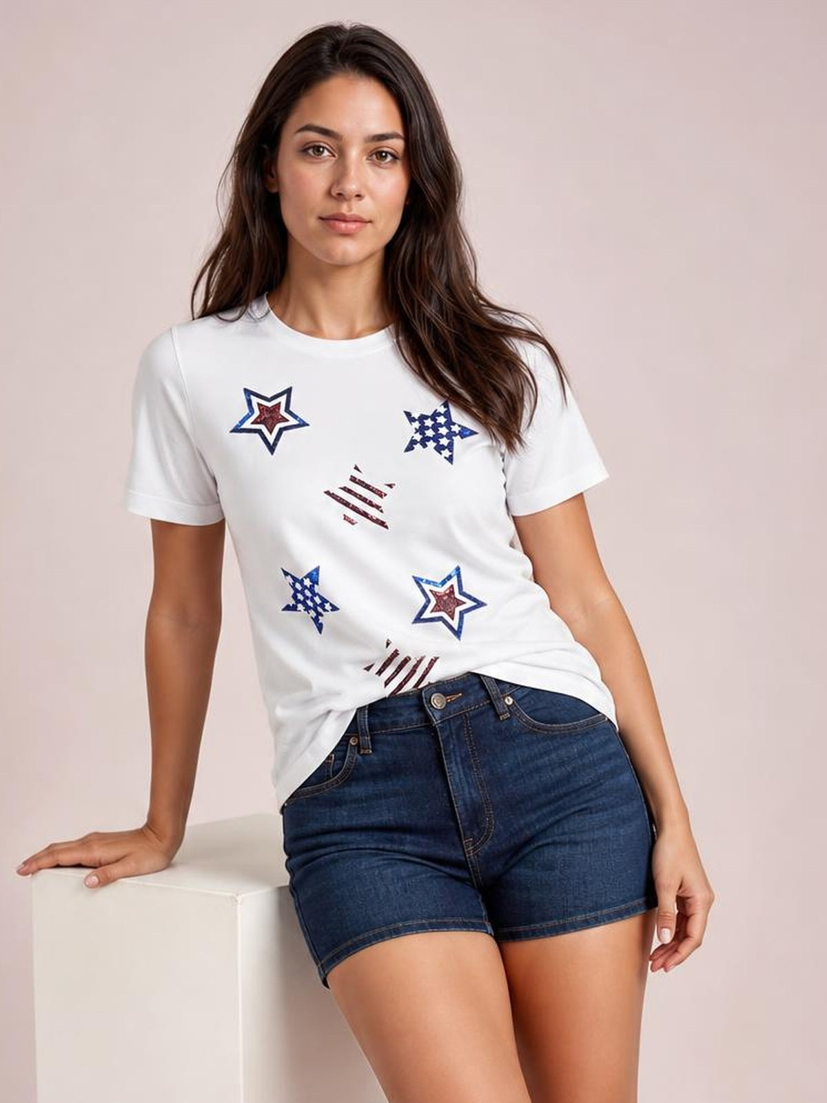 A model leans against a cube wearing a white round-neck short-sleeve T-shirt with colorful sequin star motifs and a fitted silhouette.