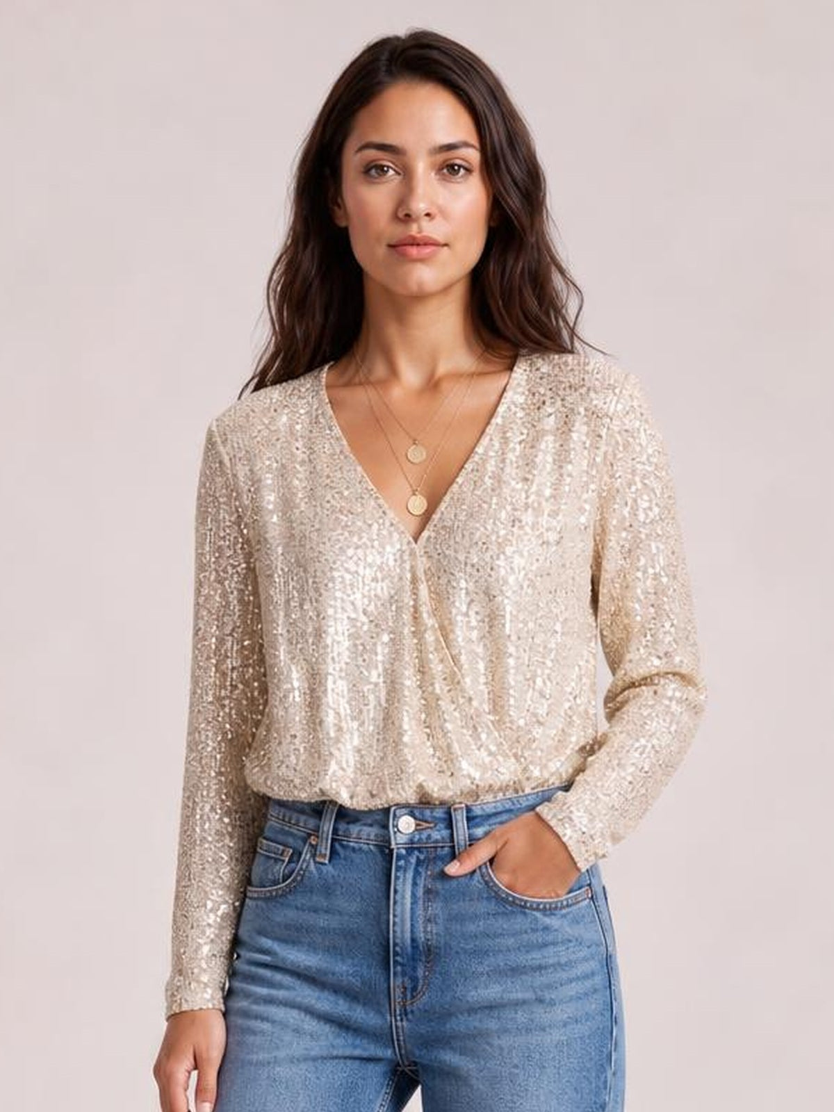 A model wears a champagne sequin surplice bodysuit with a V-neck, long sleeves, and a relaxed silhouette, posing with one hand in her jeans pocket.