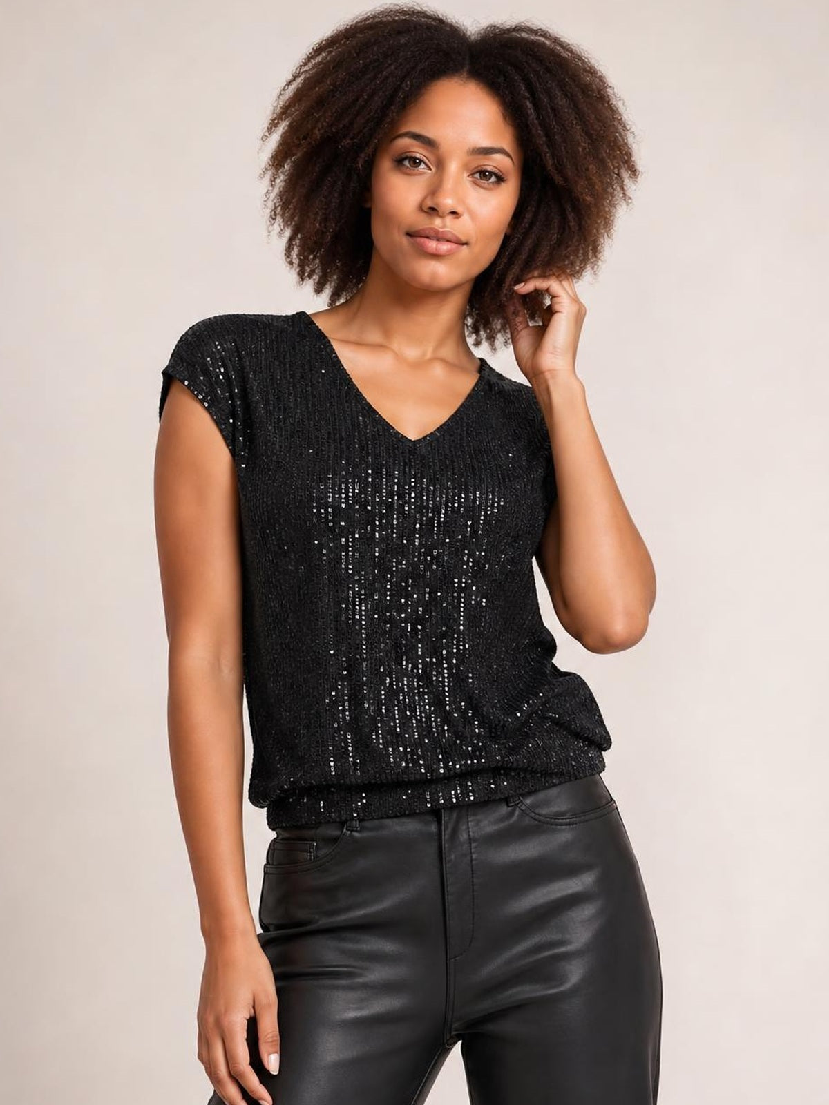 Model poses in a black sequin V-neck cap sleeve top with an opaque, relaxed silhouette and subtle drape, touching her hair with one hand.