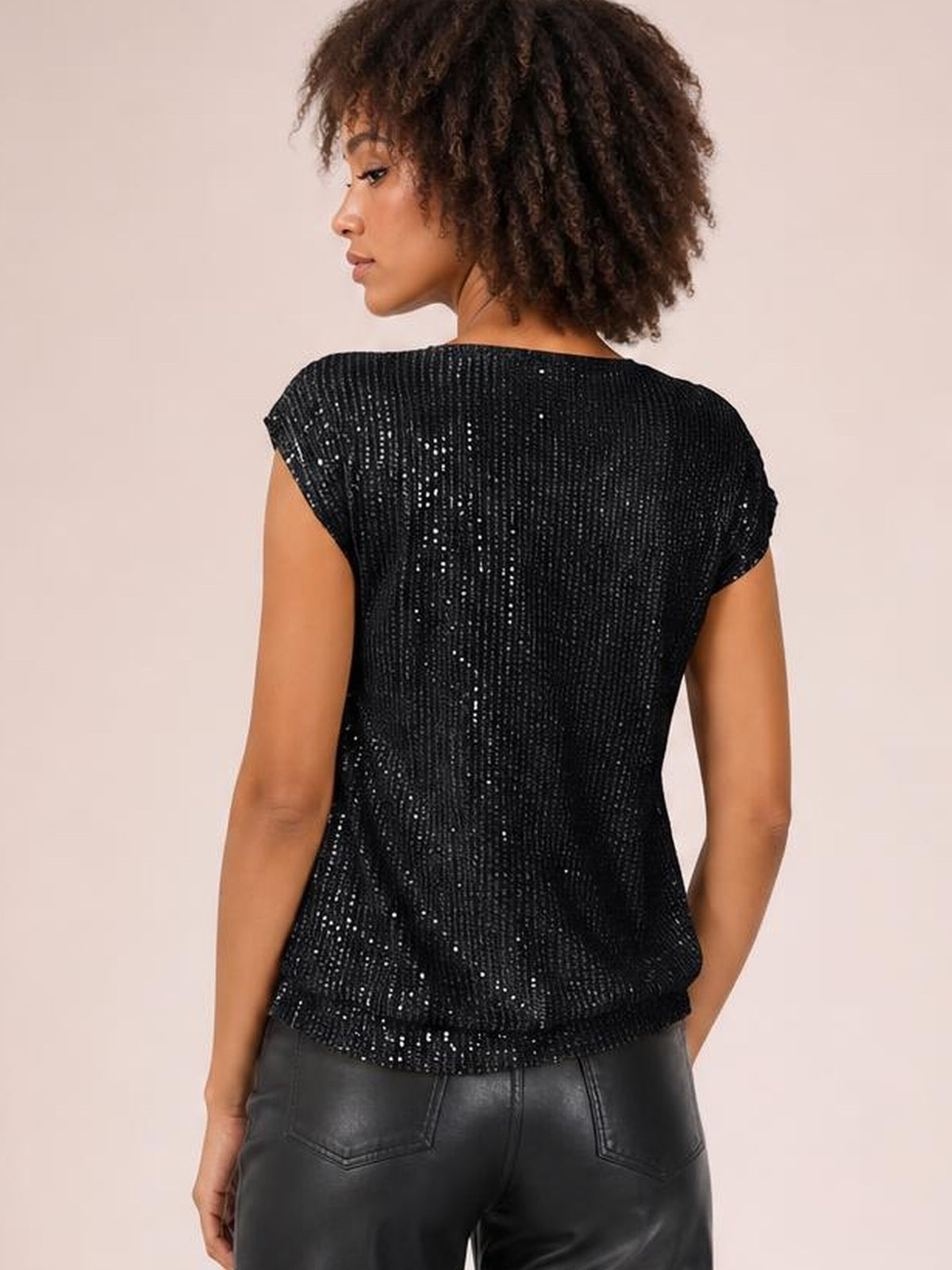 A model poses from the back wearing a black sequin top with cap sleeves, a straight silhouette, and a softly curved hem.