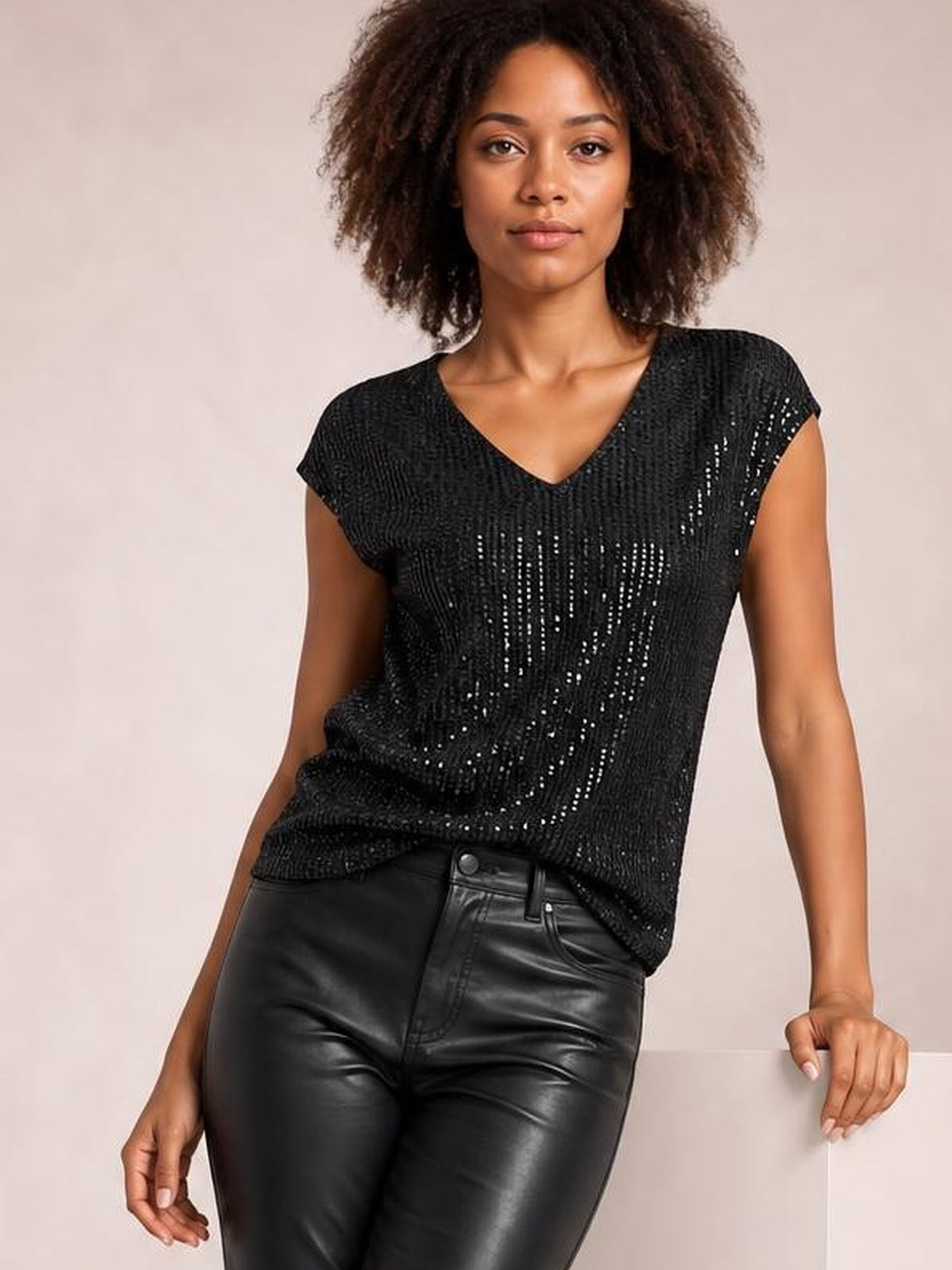 Model in a relaxed pose wears a black sequin V-neck cap sleeve top with a softly fitted silhouette and shimmering vertical texture.