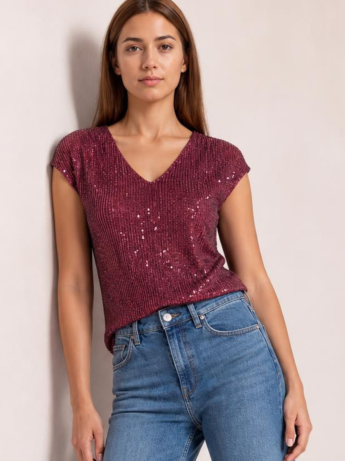 A model wears a burgundy sequin V-neck cap sleeve top with a relaxed straight silhouette and shimmering textured finish, paired with blue jeans.
