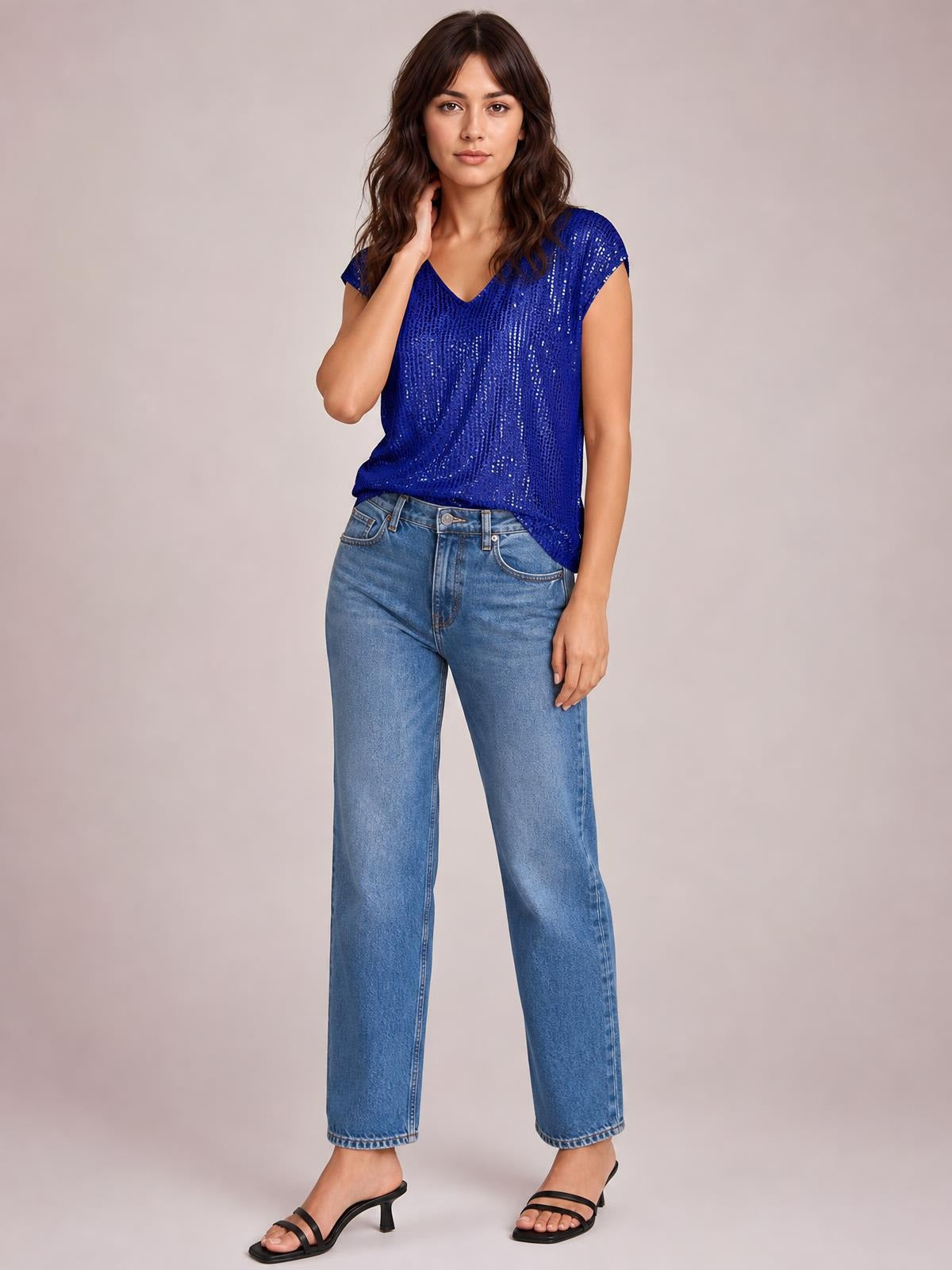 Model poses in a blue sequin V-neck cap sleeve top with a relaxed silhouette and curved hem, paired with straight-leg jeans.