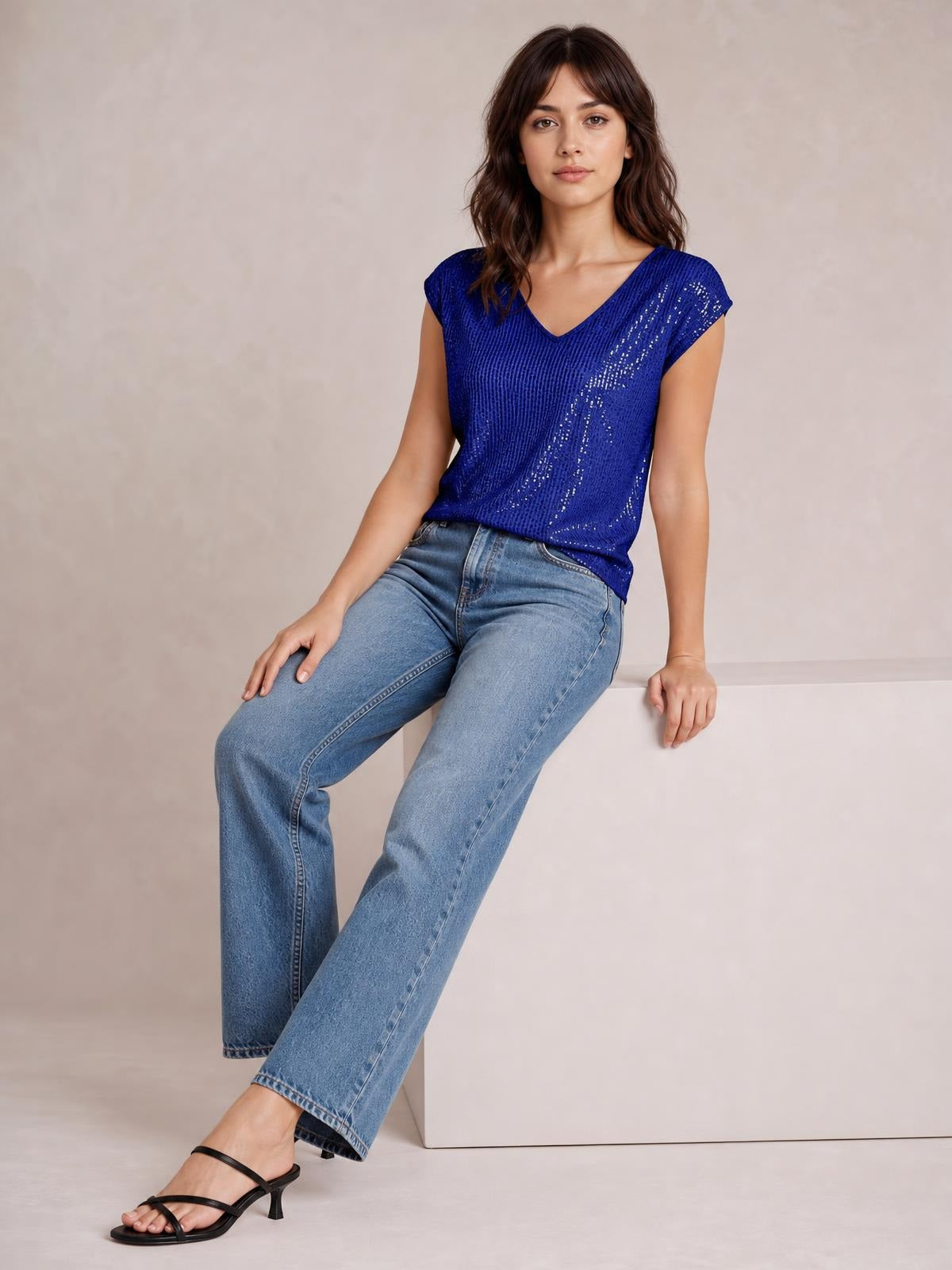 Model seated in a relaxed pose wearing a royal blue sequin V-neck cap sleeve top with a loose straight silhouette and shimmering vertical construction.
