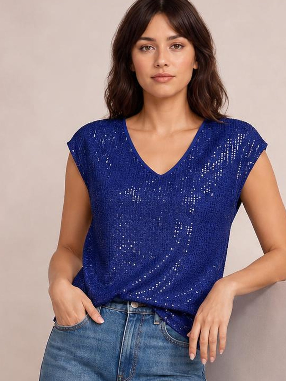 Model wearing a royal blue sequin V-neck cap sleeve top with an opaque, relaxed silhouette, posing with one hand in her jeans pocket.