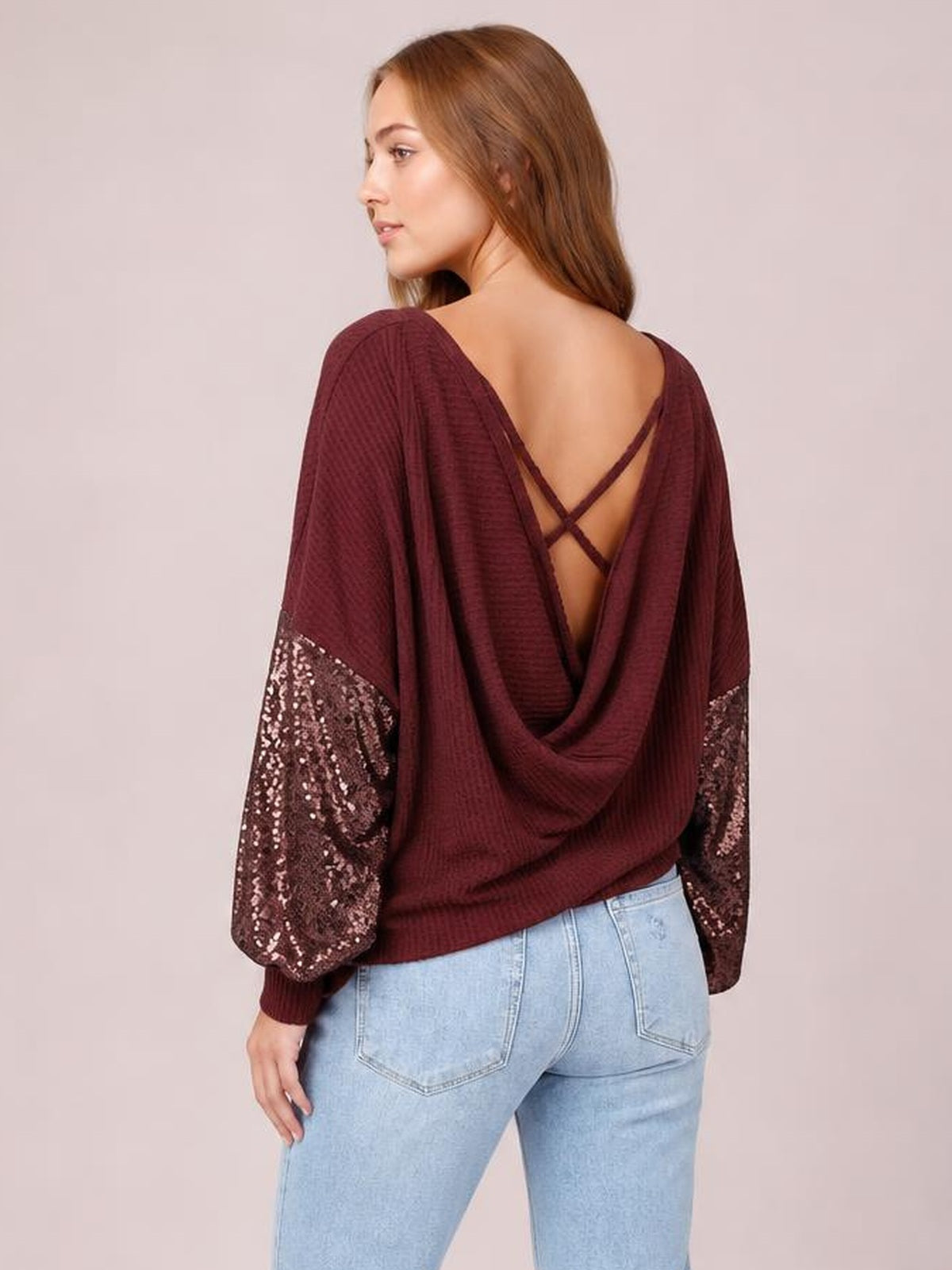 A model turns to show a burgundy sequin waffle-knit blouse with a draped open back, crisscross straps, and loose long sleeves with sequin panels.