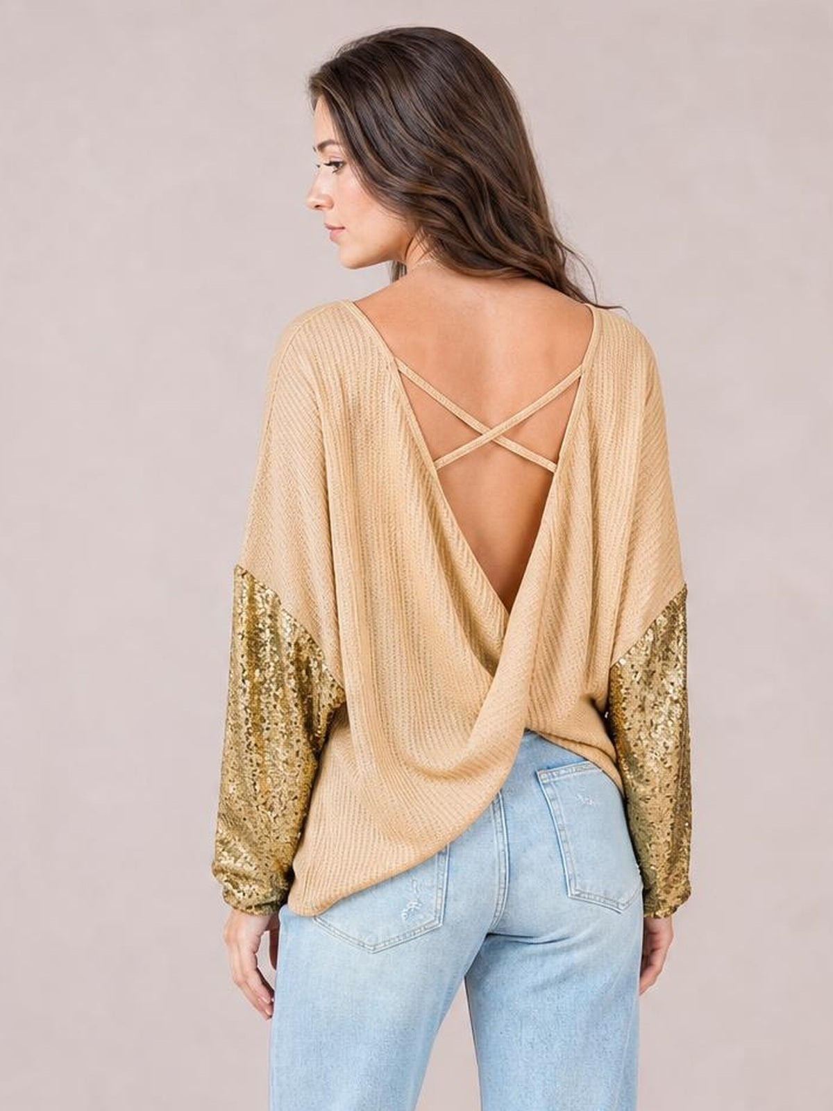 Model turned to the side shows a beige sequin waffle-knit blouse with a loose silhouette, long glittering sleeves, and an open back with crossed straps.