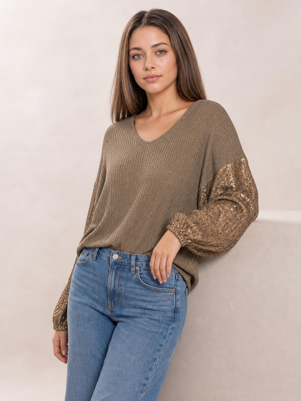 Model wears a taupe sequin waffle-knit blouse with a V-neck, relaxed silhouette, and shimmering long sleeves while leaning with one hand at her waist.