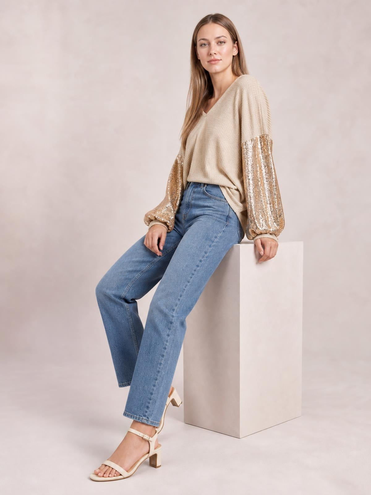 Model seated in a beige sequin waffle-knit blouse with a V-neck, relaxed silhouette, and voluminous sequin sleeves with gathered cuffs.