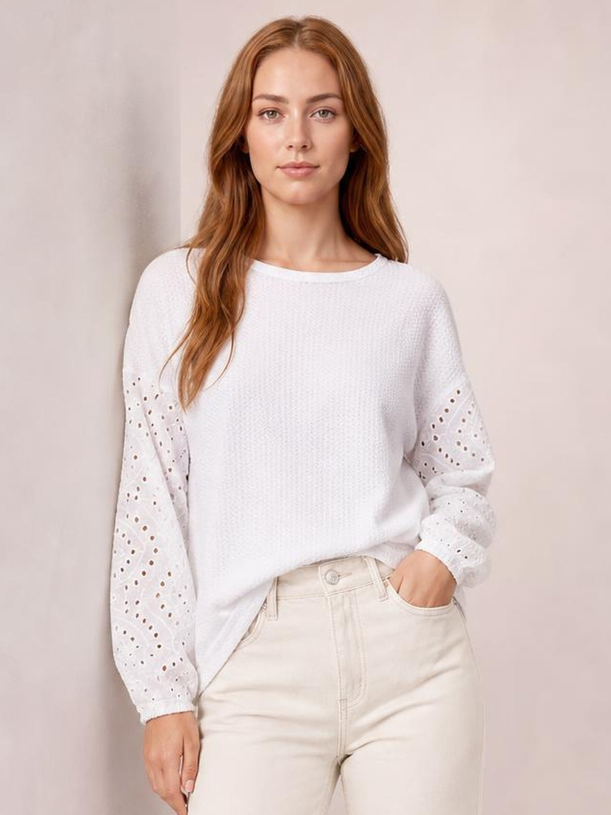 Model wears a white eyelet blouse with a relaxed dropped-shoulder silhouette, round neck, textured body, and long openwork sleeves, standing with one hand in her pocket.