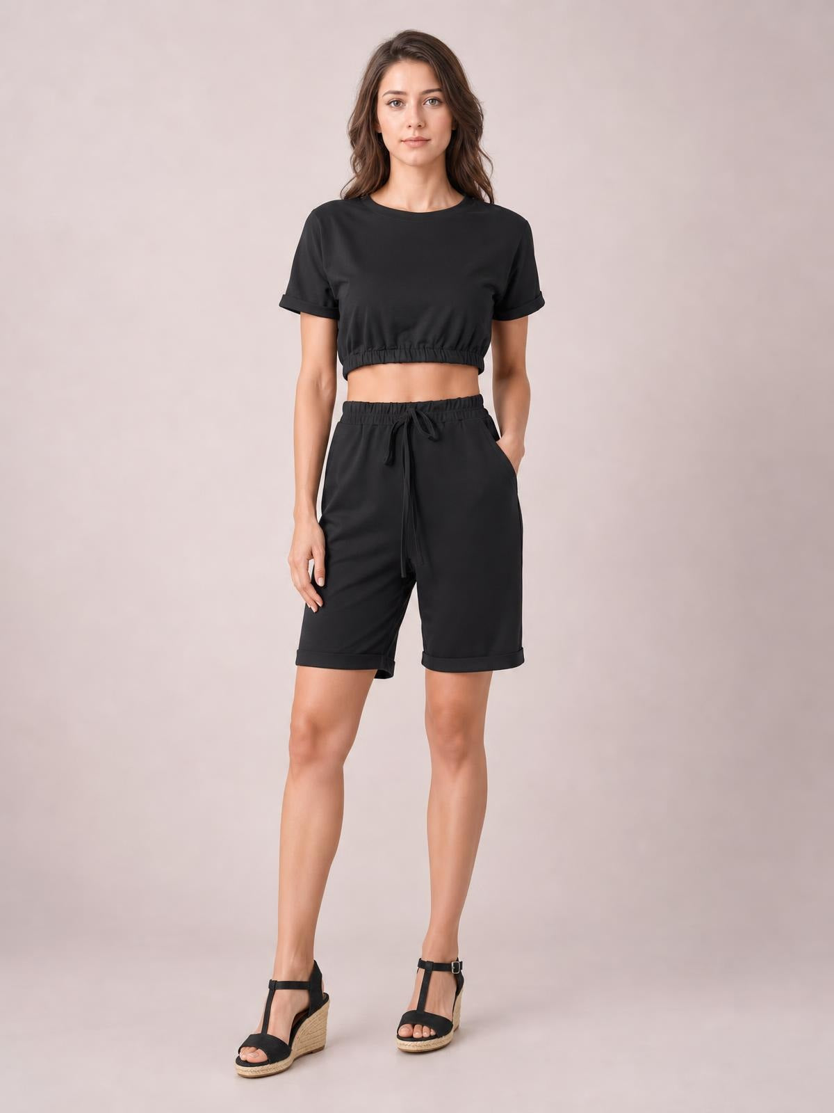 Model wearing a solid black lounge set with a cropped short-sleeve round-neck top and drawstring shorts, featuring elastic waist, tie detail, and rolled hems.