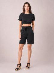 Model wearing a solid black lounge set with a cropped short-sleeve round-neck top and drawstring shorts, featuring elastic waist, tie detail, and rolled hems.