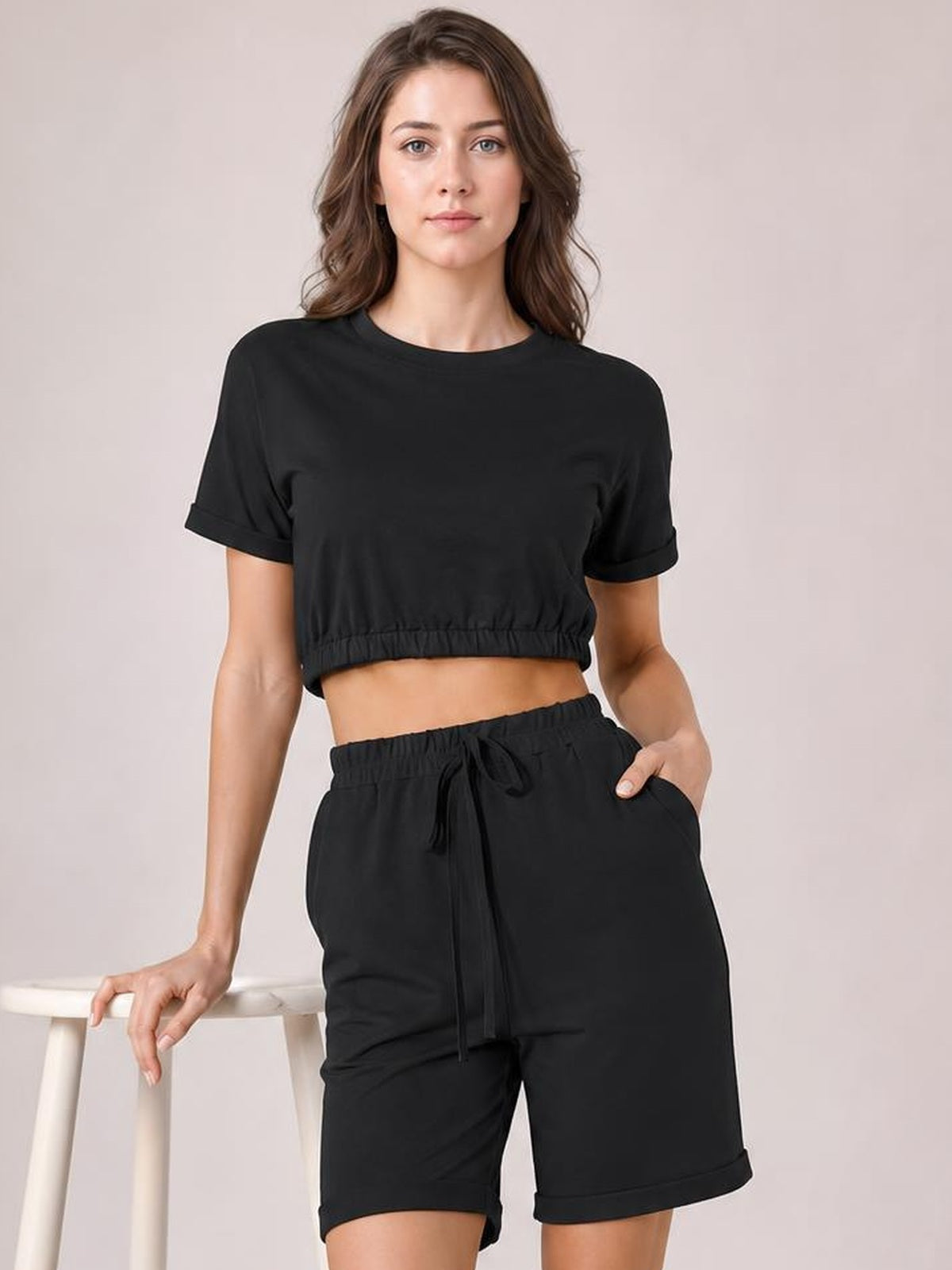 Model poses in a black minimalist lounge set with a cropped short-sleeve round-neck top and drawstring shorts featuring an elastic waist and rolled hems.