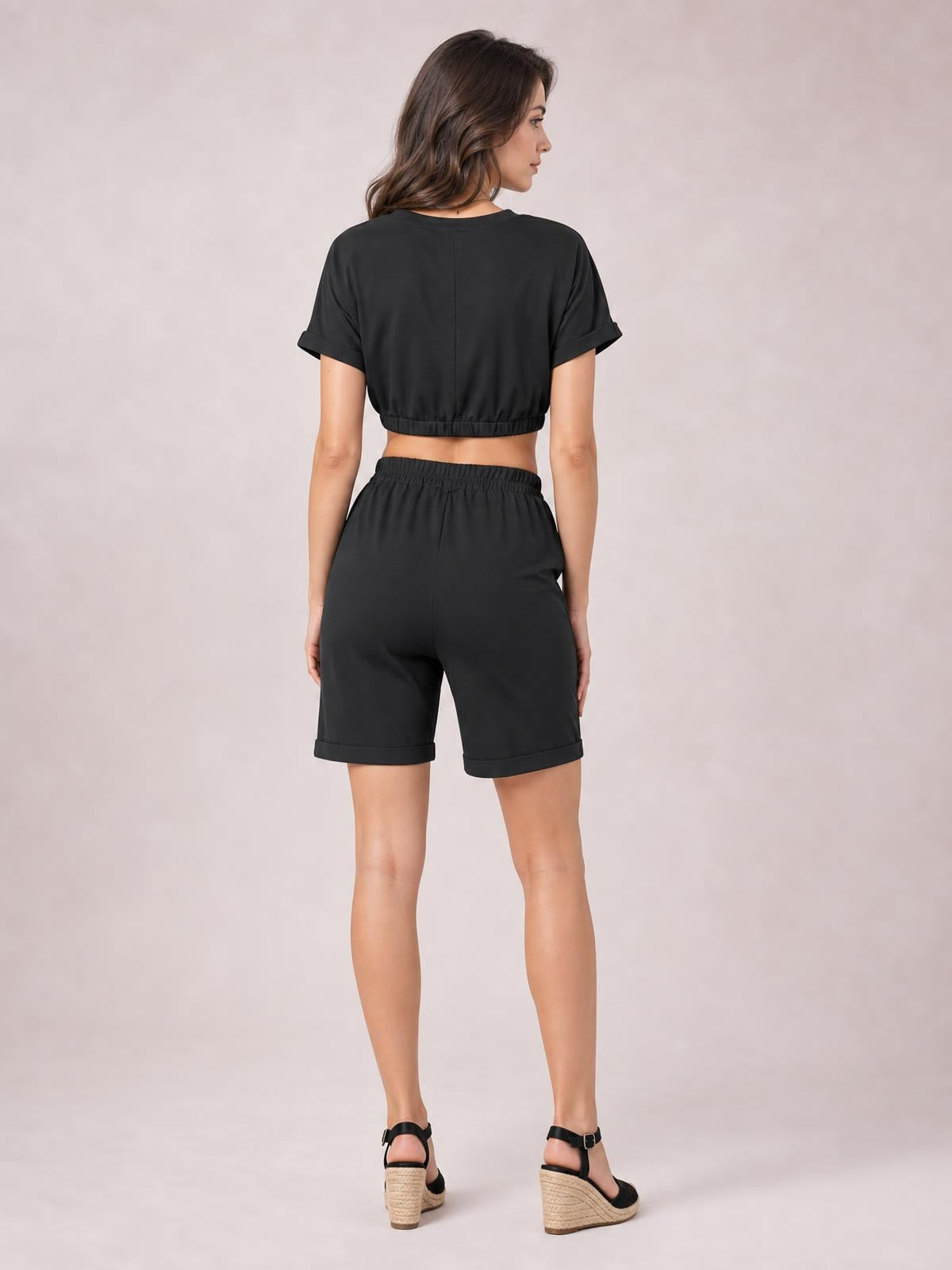 A model poses from the back in a black solid lounge set with a cropped short-sleeve top, elastic waistband, and cuffed drawstring shorts.