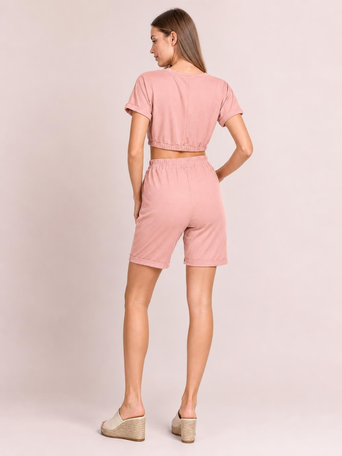 Model in a back view wears a solid pink cropped short-sleeve top with rolled cuffs and matching drawstring shorts featuring an elastic waistband and relaxed silhouette.
