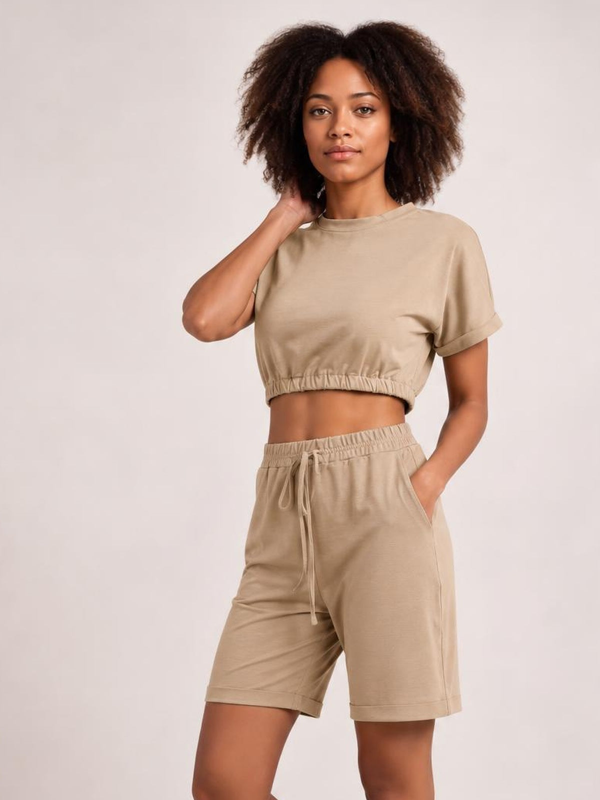Model poses in a beige lounge set with a cropped short-sleeve round-neck top, elastic hem, and drawstring shorts featuring side pockets.