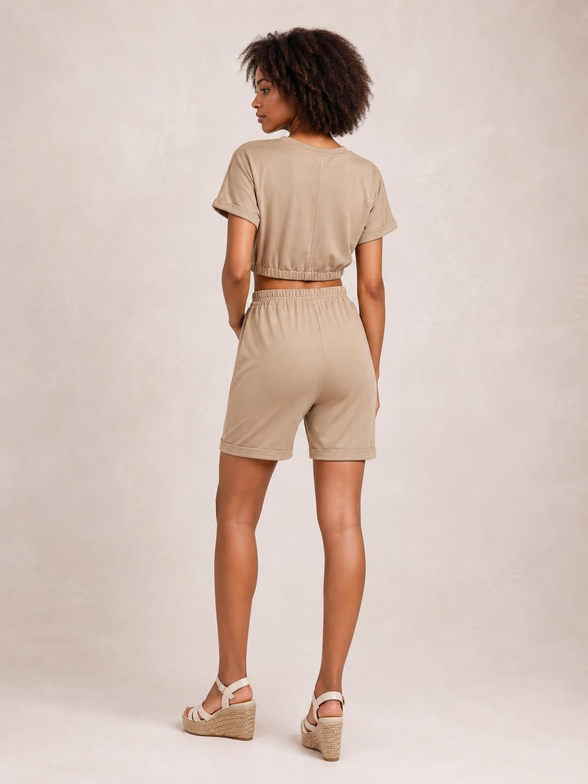 Model poses turned away in a taupe cropped short-sleeve top and matching drawstring lounge shorts with elastic waistbands and cuffed hems.