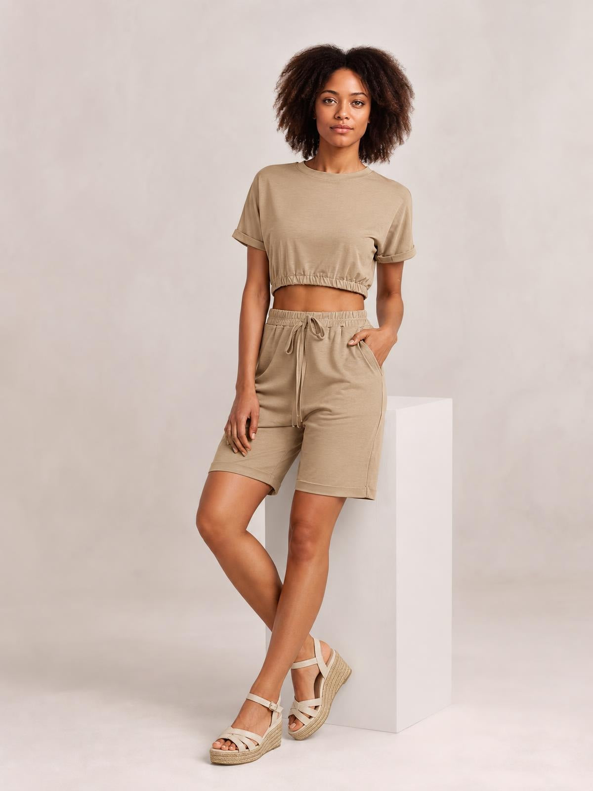 Model poses in a solid beige lounge set with a cropped round-neck short-sleeve top and knee-length drawstring shorts, featuring an elastic hem and relaxed silhouette.