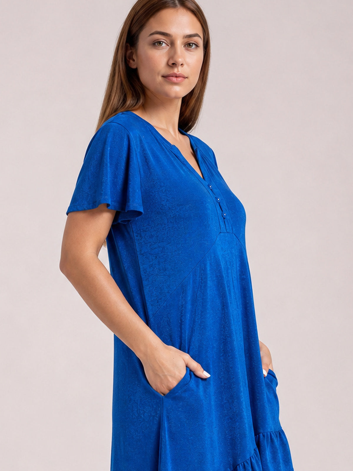 Model stands with hands in the pockets of a royal blue short sleeve dress featuring flutter sleeves, a notched neckline, relaxed silhouette, and ruffled asymmetric hem.
