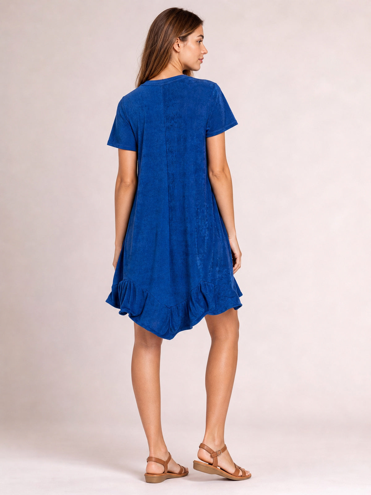 A model shows the back of a royal-blue short-sleeve dress with a relaxed silhouette, center seam, and ruffled asymmetric hem while stepping forward.