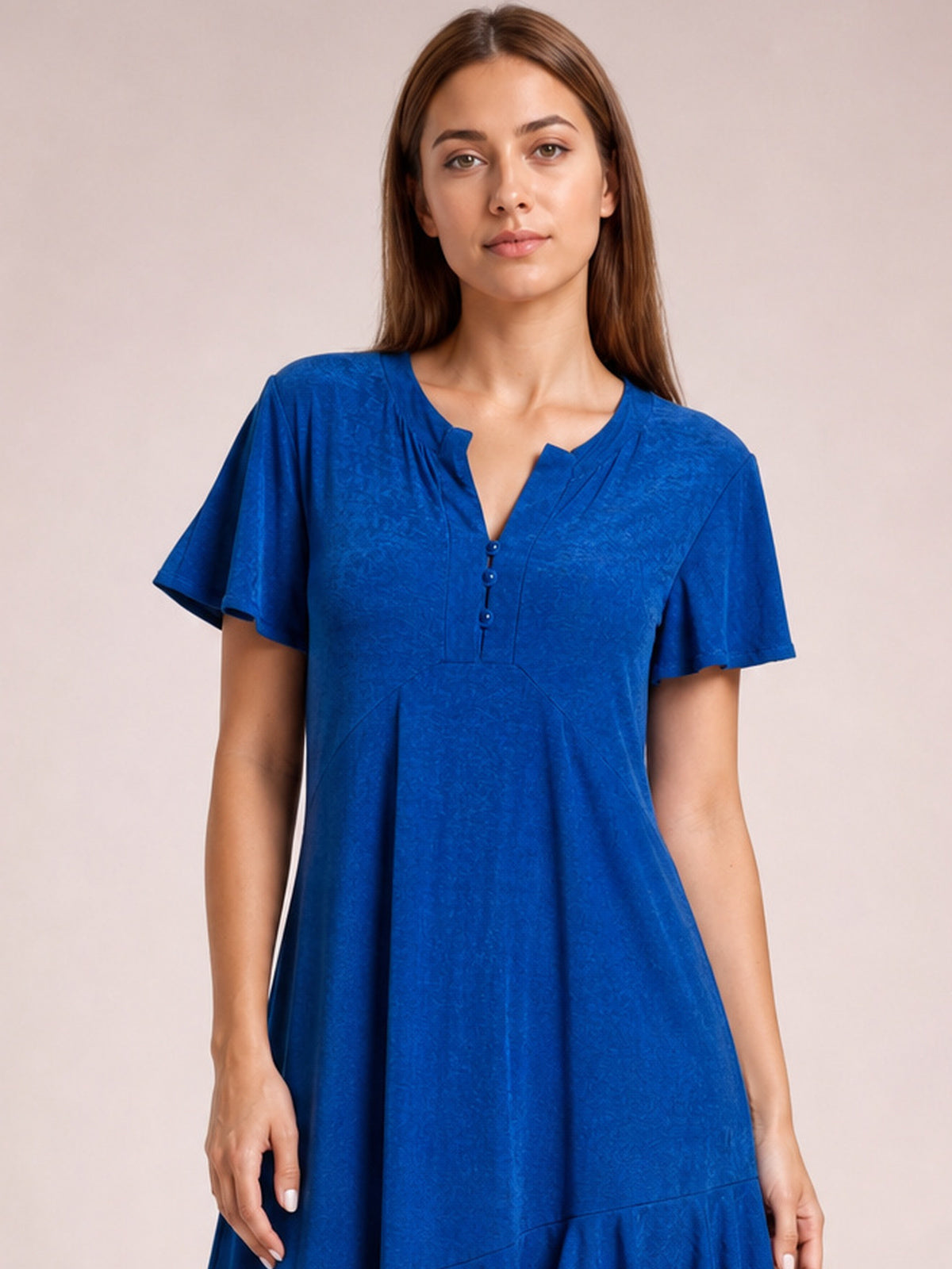 Model stands facing forward in a royal blue short sleeve dress with flutter sleeves, a notched neckline, relaxed flared silhouette, and ruffled asymmetric hem.
