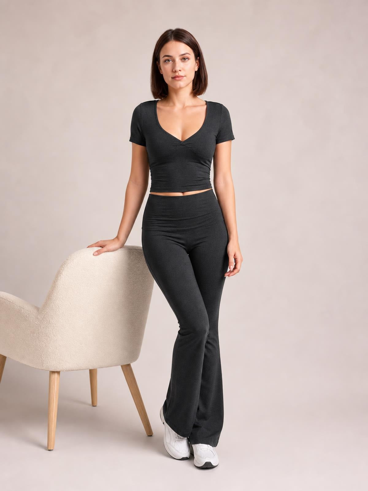 A model poses in a black short-sleeve V-neck top with a fitted cropped silhouette and high-waisted bootcut leggings, resting one hand on a chair.