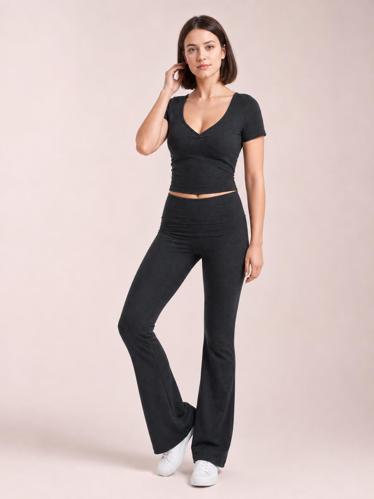 Model wears a dark charcoal short sleeve top with a V-neck crossover front and high-rise bootcut leggings, posing with one leg bent.
