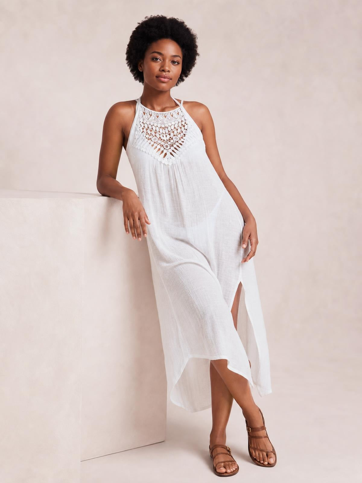 Model leans against a block wearing a white crochet cover-up dress with delicate lace detail, halter neckline, flowing silhouette, and side slit.
