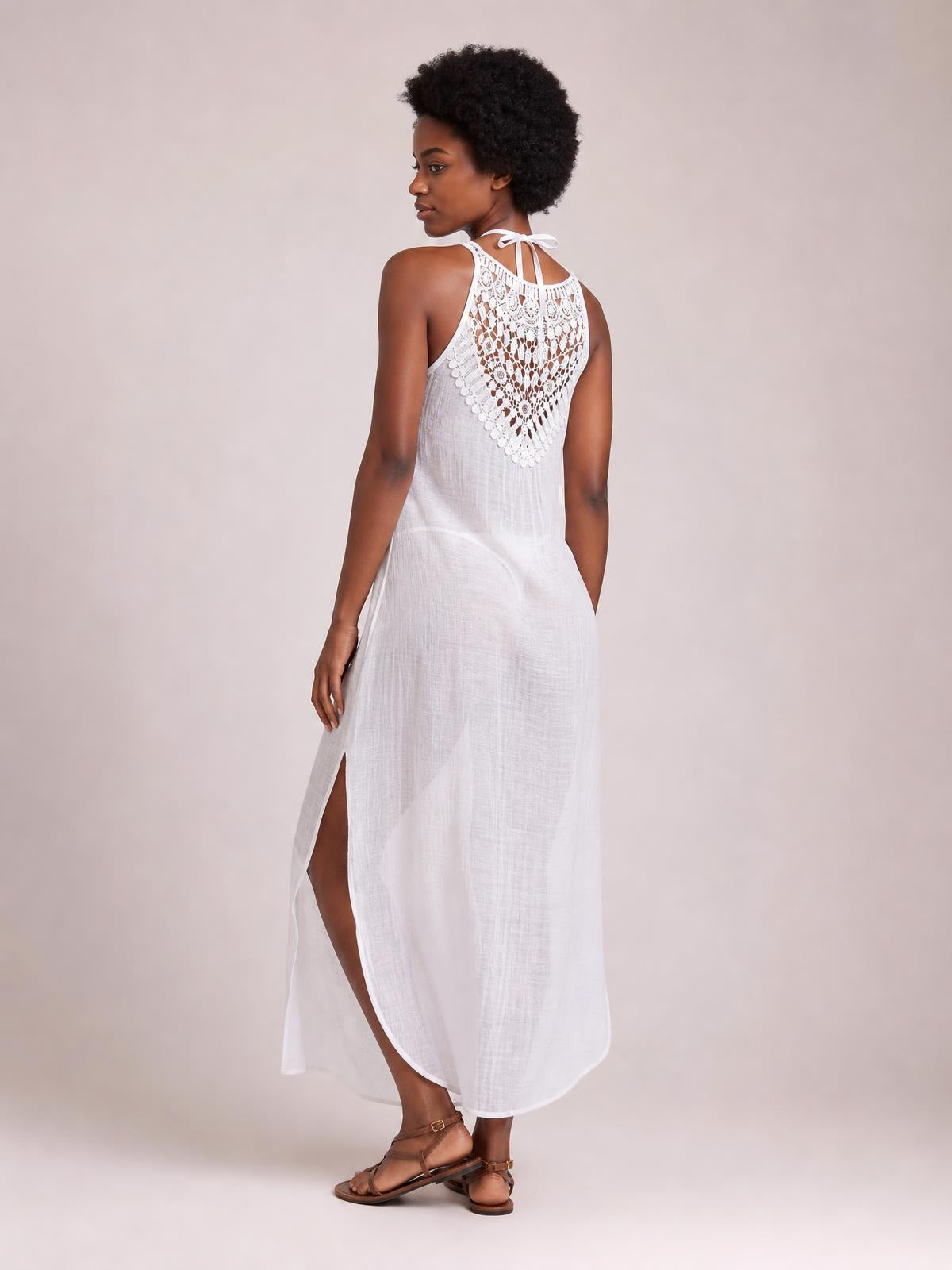 A model looks over her shoulder in a white maxi cover-up dress with a crochet lace back panel, halter tie, relaxed silhouette, and side slit.