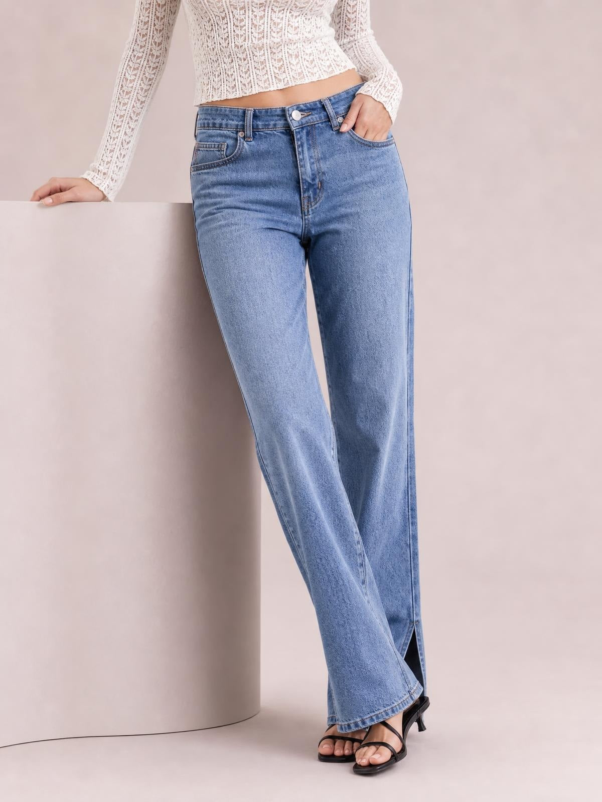 A model leans against a pedestal wearing light blue jeans with belt loops, front pockets, a button fly, straight legs, and side slits at the hems.