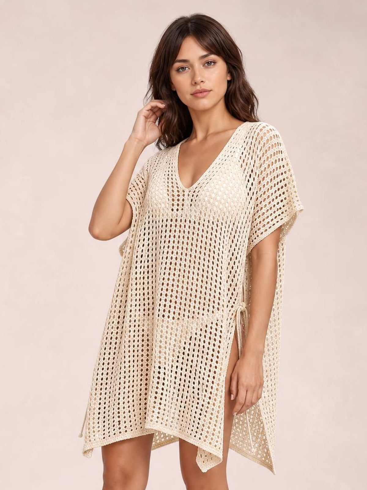Model poses in a cream side-tied V-neck cover-up with an open-knit cutout pattern, relaxed silhouette, short sleeves, and adjustable side ties.