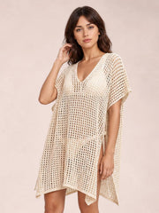 Model poses in a cream side-tied V-neck cover-up with an open-knit cutout pattern, relaxed silhouette, short sleeves, and adjustable side ties.