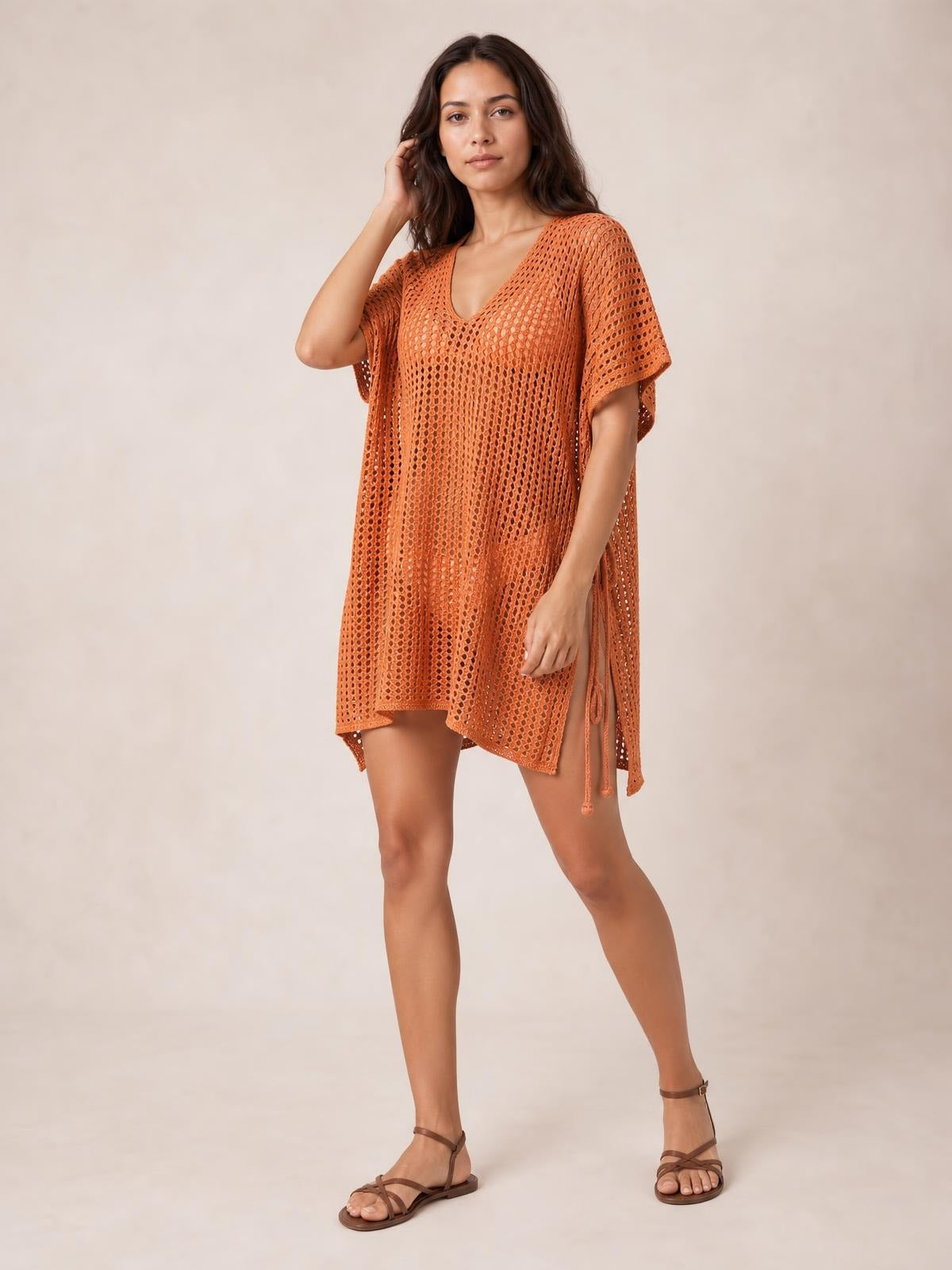 A model poses in a rust-orange V-neck cover-up with openwork cutouts, loose tunic silhouette, short sleeves, and adjustable side ties.
