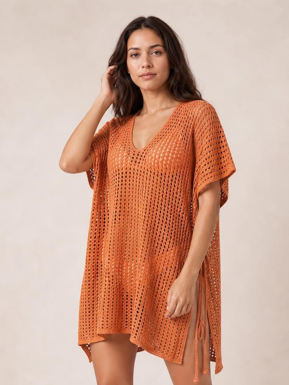 Model touches her hair while wearing a rust-orange side-tied V-neck cover-up with a loose silhouette, short sleeves, open-knit cutouts, and side ties.