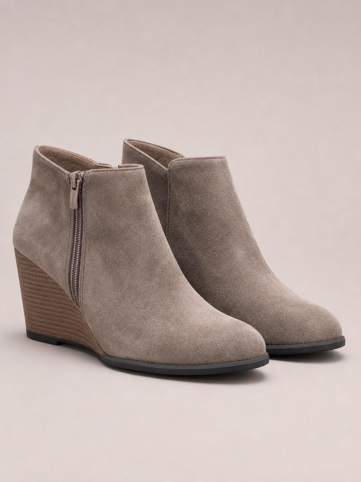Taupe suede wedge boots feature high heels, side zippers, smooth ankle-height uppers, and dark rubber soles with a sleek tapered silhouette.