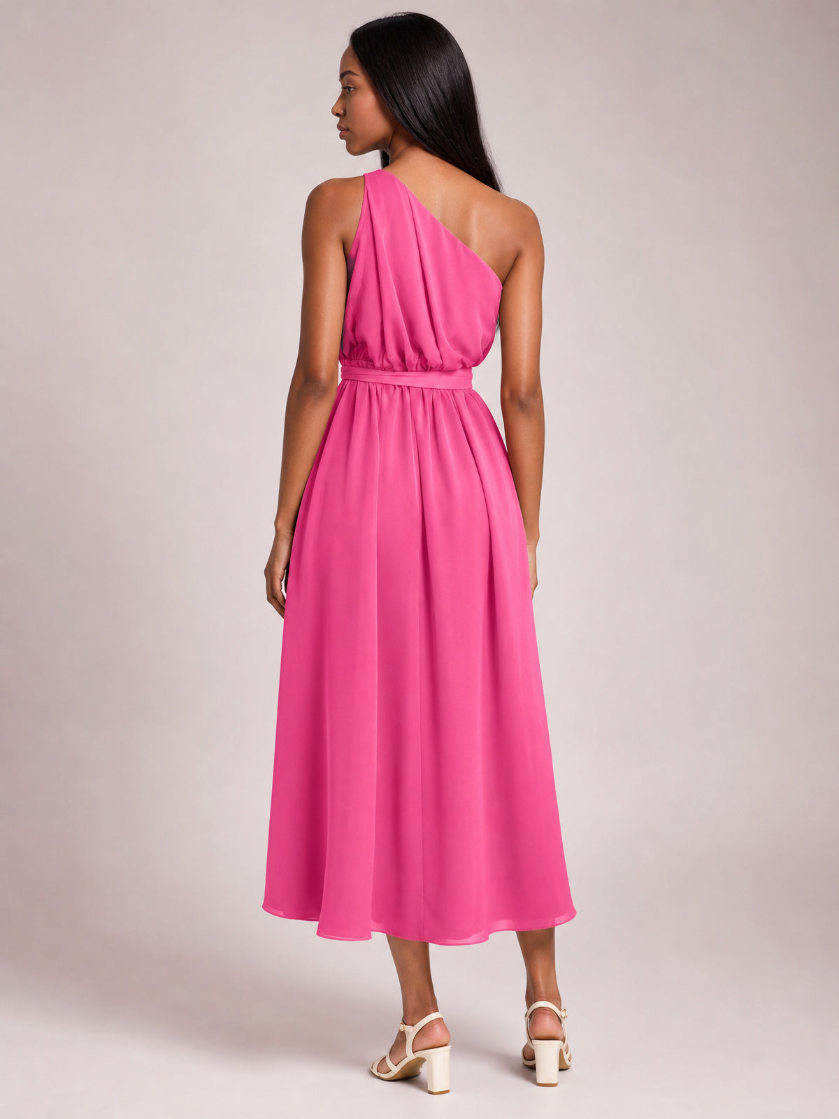 Model shown from behind in a bright pink single shoulder midi dress with a gathered bodice, defined waist belt, and flowing, softly draped skirt.