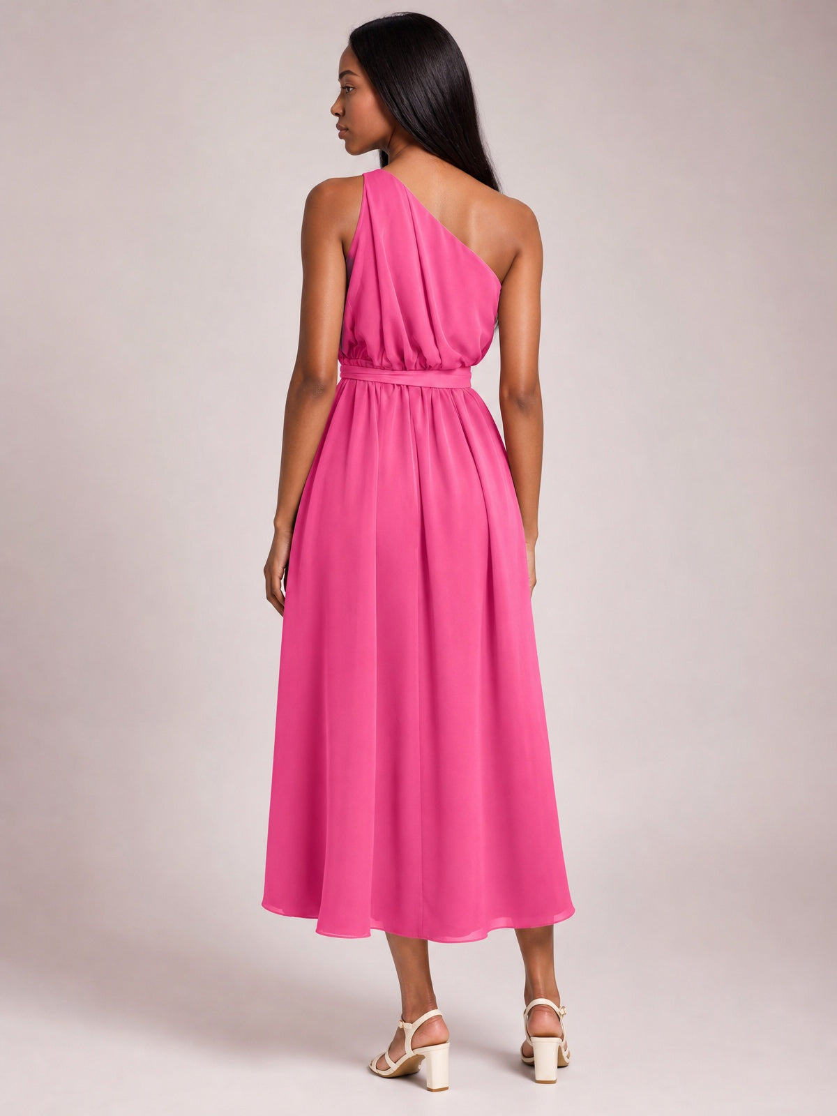 Model shown from behind in a bright pink single shoulder midi dress with a gathered bodice, defined waist belt, and flowing, softly draped skirt.