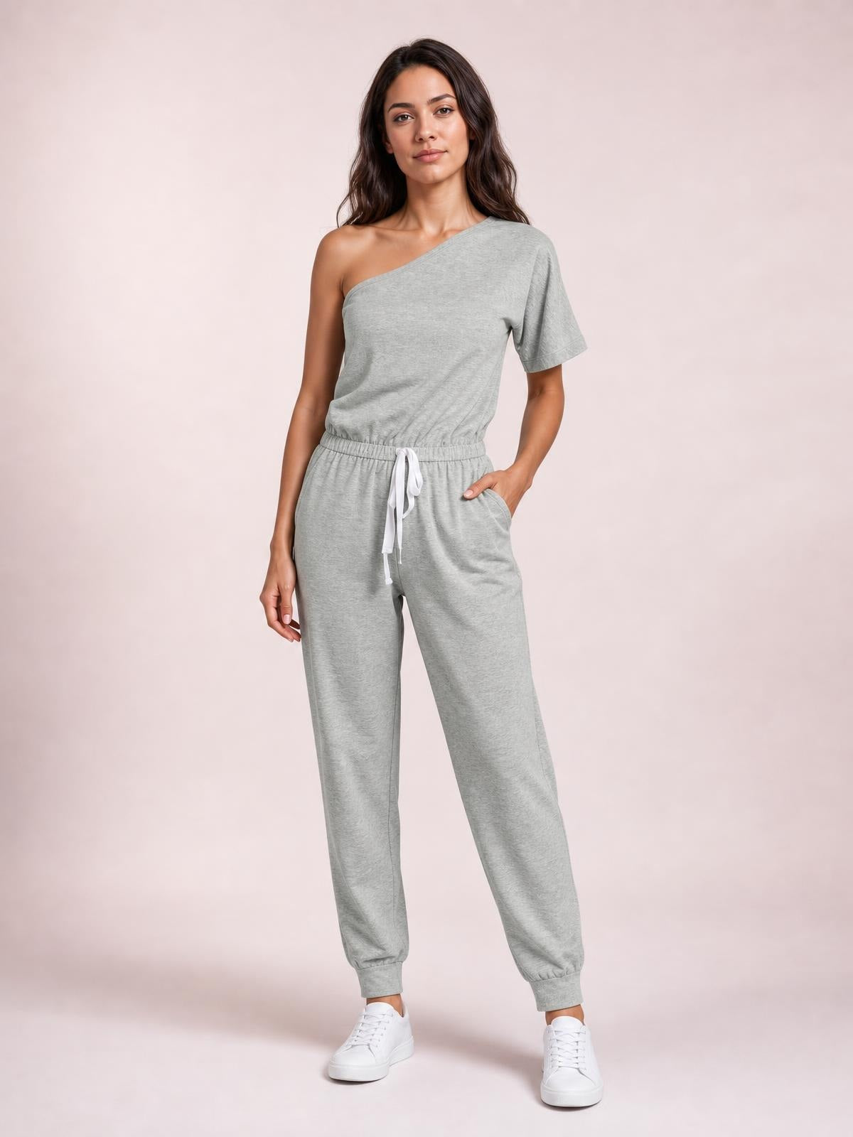 Model wearing a light gray single-shoulder short-sleeve jumpsuit with a tied drawstring waist, relaxed tapered legs, and one hand in a pocket.