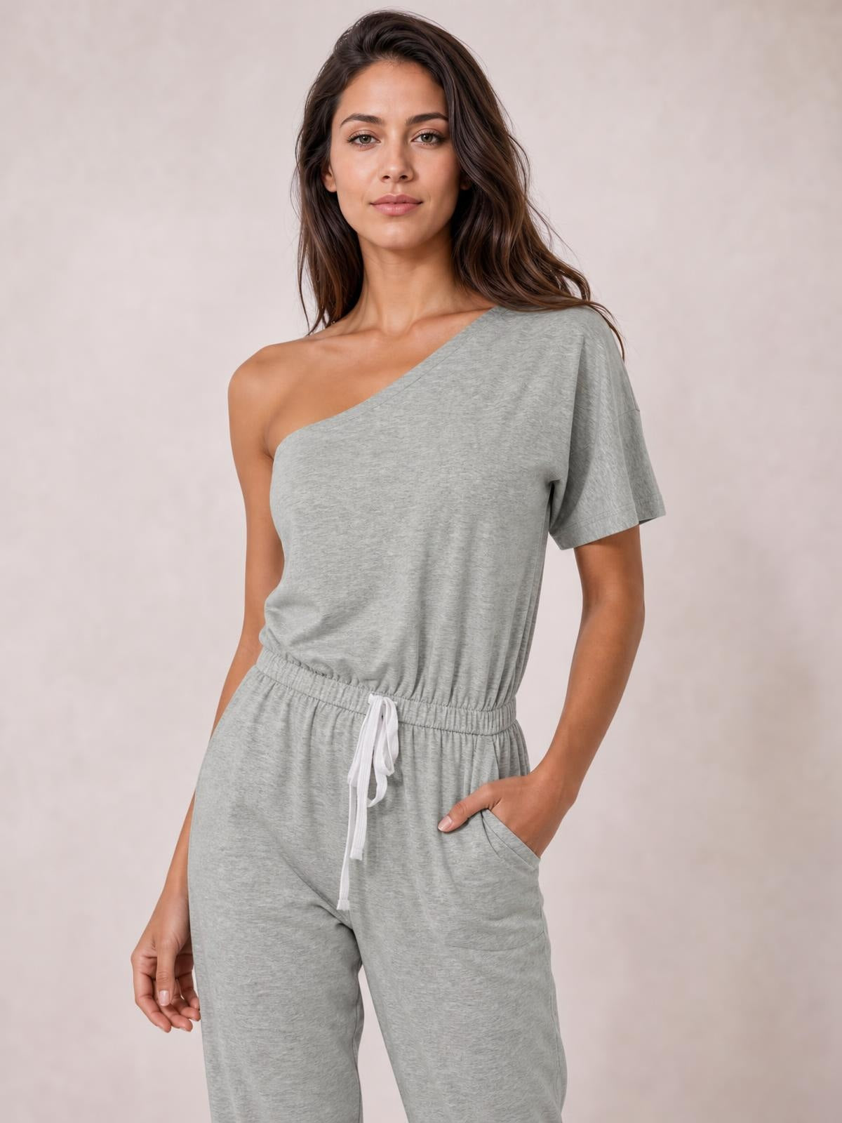 Model wearing a light gray single-shoulder short-sleeve jumpsuit with a relaxed silhouette, elastic waist, tied drawstring, and side pockets.